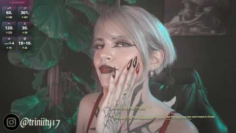 Media: Video of a young woman with short, platinum blonde hair, wearing dark lipstick, and a black choker. She has intricate black tattoos on her fingers and neck. The background features a green plant and dim lighting.