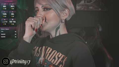 Media: Video of a pale-skinned woman with short platinum blonde hair, wearing a black \"Mot\u00f6rhead\" t-shirt, holding a microphone. Background shows dimly lit, green screen, with social media and streaming stats.