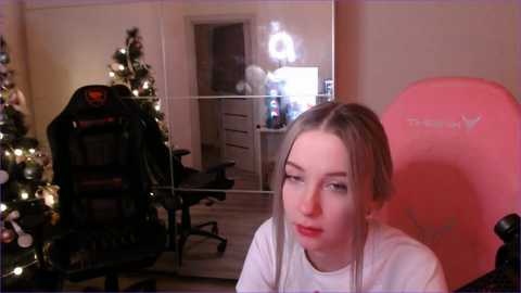 Video of a young woman with straight blonde hair, wearing a white shirt, sitting in a pink gaming chair, in a cozy, Christmas-decorated room with a black gaming chair and a lit tree.