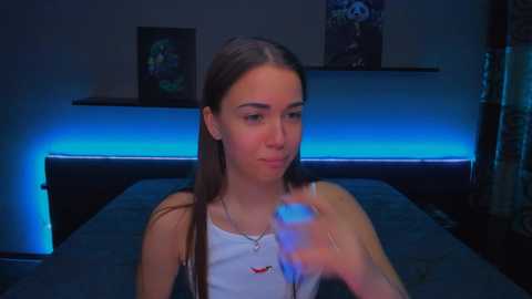 Media: Video of a young Caucasian woman with straight brown hair, wearing a white tank top, sitting on a dark bed. She's holding a glowing blue device. The room is dimly lit with blue LED lights on the wall.