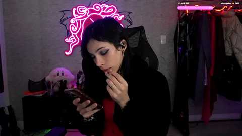 Media: Video of a young woman with long black hair and dark makeup, sitting in a dimly lit room with a neon pink skull wall decal, holding a phone, wearing a black jacket.