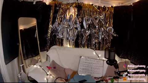 Media: Video of a dimly lit, festive room with metallic \"Happy New Year\" balloons, string lights, a black microphone stand, and a white sheet covering a table.