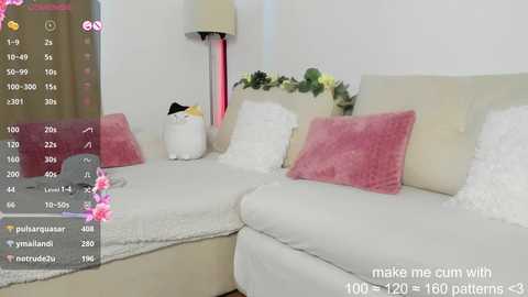 Media: Video of a modern living room featuring a white sectional sofa adorned with fluffy pink and white pillows, a white rug, and a tall floor lamp. A small plant sits on a white table in the background.