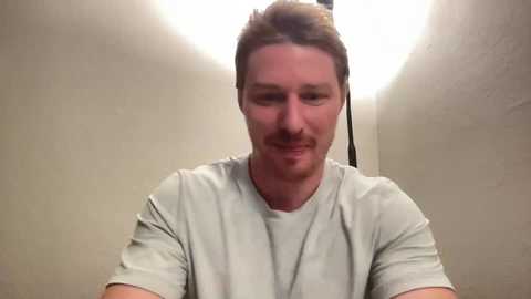 Media: Video of a smiling white man with short brown hair, wearing a light gray T-shirt, standing in a narrow, dimly lit, beige-walled corridor.