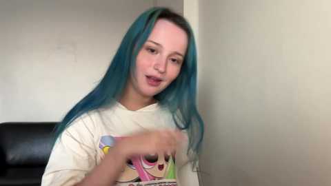 Media: A video of a young woman with long, vibrant teal hair, wearing a white T-shirt featuring colorful cartoon characters. She is indoors, with a plain white wall and a black leather couch in the background.