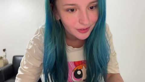 Media: Video of a young woman with long, vibrant blue hair, fair skin, and a white T-shirt featuring a large, colorful anime eye graphic. She smiles softly, set against a plain white background.