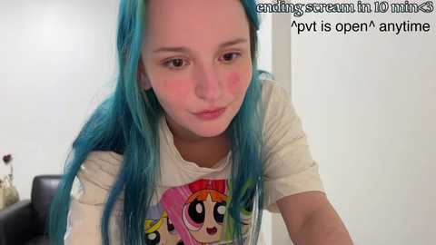 Media: Video of a young woman with long, blue hair, wearing a graphic tee, in a livestream setting.