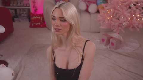 Media: Video of a blonde woman in a black tank top, kneeling on a carpeted floor, with a pink Christmas tree and wrapped gifts in the background.