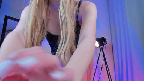 Media: Video of a blonde woman with long hair, wearing a black lace top, leaning over a desk with a microphone stand and light.