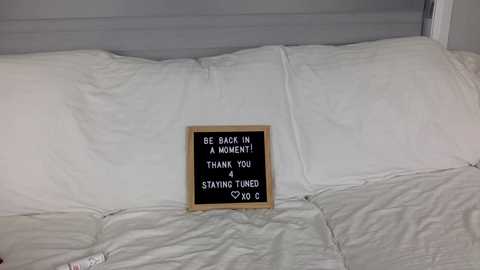 Media: Video of a neatly made bed with white sheets and a wooden-framed black chalkboard sign reading, \"Be back in 4 hours. Thank you. Stay tuned.\