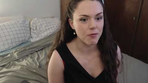 Media: Video of a young woman with long brown hair, wearing a black sleeveless dress, sitting on a bed with white and gray patterned pillows, in a dimly lit bedroom.