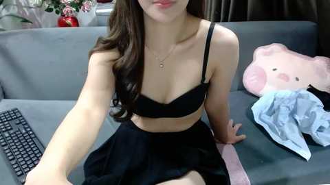 Media: Video of an Asian woman with long brown hair, wearing a black bra and skirt, sitting on a gray sofa, with a keyboard, pink plush toy, and clothes beside her.