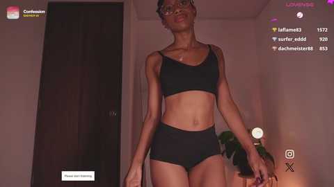 Media: Video of a slender, dark-skinned woman with glasses in a black sports bra and shorts, standing in a dimly-lit room with a closed door and a plant.