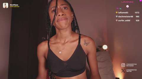 Media: Video of a Black woman with braided hair, wearing a black bralette, standing indoors. She has a tattoo on her shoulder and a small necklace. Background is dimly lit, with a \"Lovemeet\" watermark.