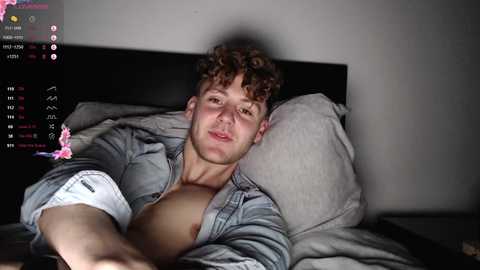 Media: A video of a shirtless, muscular, young man with curly hair and fair skin, lying on a bed, wearing a light blue denim jacket, in a dimly lit room.