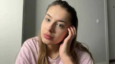 Media: Video of a young Caucasian woman with light skin, green eyes, and long, straight blonde hair, wearing a light pink t-shirt, leaning on her elbow in a plain, gray-walled room.