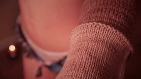 Media: A close-up video of a person's arm in a beige, knitted sweater, with a blurred background featuring a lit candle and a partially visible person in a black top. The image focuses on the texture of the sweater.