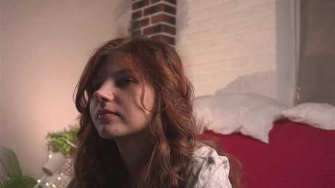 Media: Video of a young woman with wavy, light brown hair, wearing a light gray blouse, sitting on a red sofa in a dimly lit room with white brick walls and a potted plant in the background.