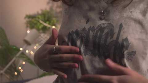 Media: Video of a woman with light skin, wearing a grey t-shirt with a black snake tattoo, applying henna to her chest, surrounded by fairy lights and a green plant.