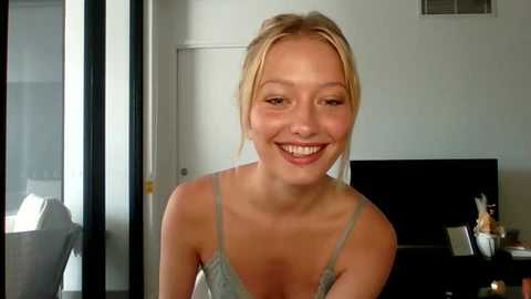 Media: Video of a smiling, blonde Caucasian woman with fair skin in a light green lace camisole, sitting indoors near a window with a TV and decor in the background.