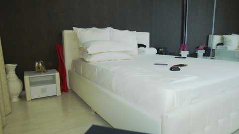 Media: Video of a minimalist bedroom with a white bed, white pillows, and white sheets. Dark walls, red vase, and a black chair.