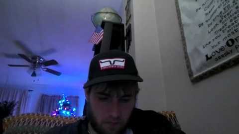 Media: Video of a young man in a dark hat, looking down, in a cozy room with a Christmas tree, a ceiling fan, and a motivational wall quote.