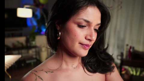 Media: Video of a light-skinned woman with shoulder-length black hair, wearing minimal makeup, smiling softly. She has a small tattoo on her shoulder. The background is a dimly lit living room with blurred decor, including a lamp and plants.