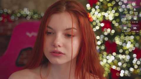 Media: Video of a young woman with long, straight red hair, fair skin, and a slight nose stud. She wears a neutral expression, standing in front of a Christmas tree with colorful lights and decorations.