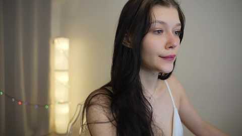 Media: Video of a young, fair-skinned woman with long, dark brown hair, wearing a white spaghetti-strap top, standing indoors against a softly lit background with a vertical light fixture.