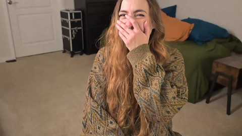 Media: Video of a young woman with long, wavy brown hair, laughing and covering her mouth, wearing a patterned sweater, sitting on a bed with green and blue sheets, in a beige-carpeted room with white walls and a black dresser.