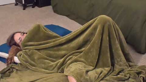 Media: Video of a woman with long brown hair wrapped in a green blanket, lying on a beige carpet, with a blue pillow and a dark green blanket beside her.