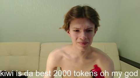 Media: Video of a young, pale-skinned woman with short brown hair, topless, sitting on a beige couch against a plain light green wall. Text overlays: \"Kwii is da best 2000 takes on my god\