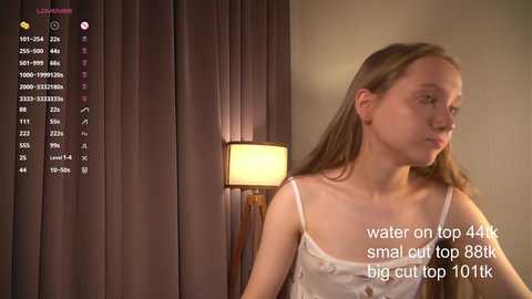 Media: A video of a young Caucasian woman with fair skin and long brown hair, wearing a white camisole, standing in a dimly-lit room with a lamp and curtains. Text overlays detail water consumption and a small cut.