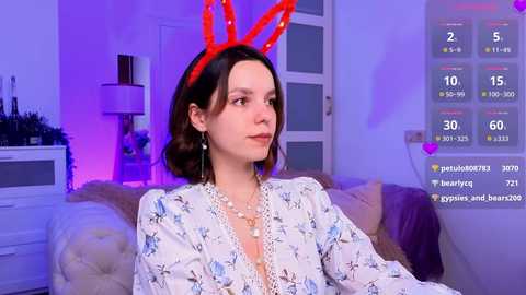 Media: Video of a young woman with fair skin, dark hair, wearing a red bunny headband, white floral dress, and silver necklace, sitting on a beige couch in a purple-lit room.