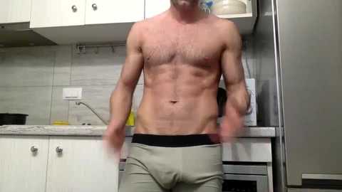 Media: Video of a shirtless, fit man with a trimmed chest and light skin, wearing beige boxers, standing in a modern kitchen with white cabinets, gray backsplash, and stainless steel appliances.