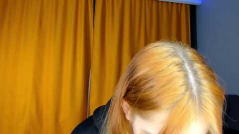 Media: Video of a person with shoulder-length, light orange hair, viewed from the back, against a backdrop of mustard-yellow curtains. The person is wearing a dark top, and the background is slightly blurred.