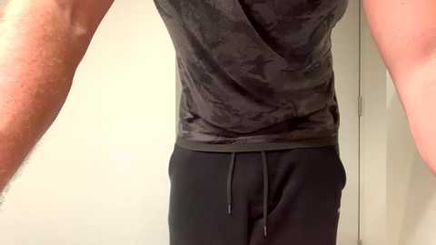 Media: Video of a person wearing a black, sleeveless, patterned tank top and black sweatpants, with a beige wall and slightly blurred arms visible in the frame.