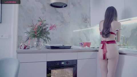 Media: Video of a woman with long brown hair, wearing a red apron, standing in a modern, white kitchen with marble backsplash, cooking utensils, and a potted plant.