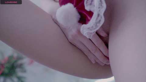 Media: Video of a woman's lower body, wearing white lace panties with red trim and a fluffy pom-pom, holding her genitals. Background is blurred with soft lighting.