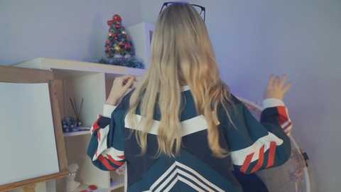 A video of a blonde woman in a dark green and white striped sweater, facing away, hands on hips, in a brightly lit room with a Christmas tree and shelves visible in the background.