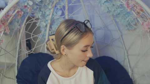 Media: A video of a young woman with light brown hair in a bun, wearing glasses, a white shirt, and a navy cardigan, sitting in a white hanging chair with floral accents, indoors.