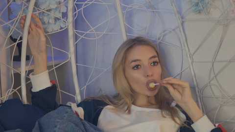 Media: A video of a blonde woman in a white shirt, eating a lollipop, lying in a white, wicker chair surrounded by decorative branches.