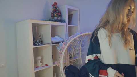 Video of a young woman with long, blonde hair wearing a white and black sports jersey, standing beside a white shelving unit with a decorative ceramic chicken on top, in a cozy room with a hanging egg chair.