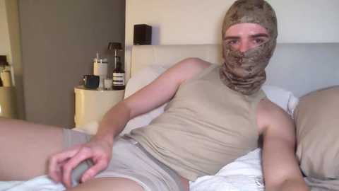 Video of a young man with fair skin, wearing a beige tank top, gray shorts, and a beige bandana covering his face, lying on a bed with white sheets, in a dimly lit room.