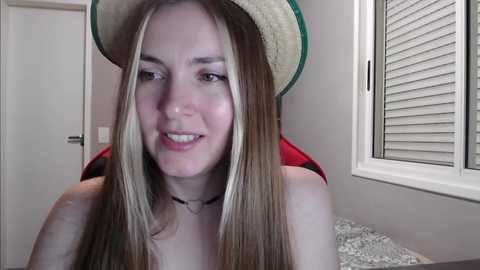 Media: Video of a smiling, fair-skinned woman with long, straight blonde hair and a straw hat, wearing a red top, sitting indoors with white blinds in the background.