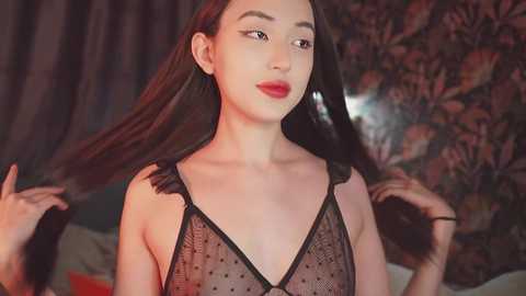 Media: Video of a young woman with long, dark hair, fair skin, and red lipstick, wearing a black, sheer polka-dot lingerie top, holding her hair. Background features a floral-patterned wall.