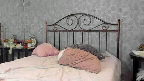 Media: Video of a messy bedroom with a wrought-iron bed, pink and grey bedding, grey hexagonal wallpaper, and cluttered nightstands.