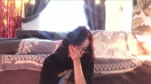 Media: Video of a woman with long dark hair, wearing glasses, and a black t-shirt, sitting on a sofa with patterned cushions, in a dimly lit room with a red and gold lamp and a window with closed curtains.