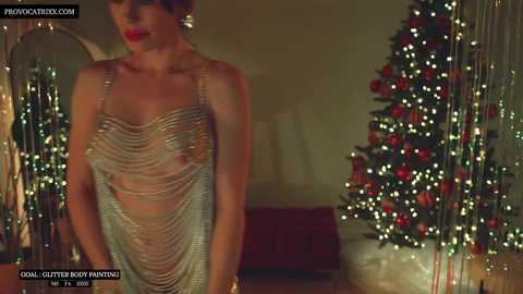 Media: Video of a woman with short brown hair, wearing a sheer metallic dress, standing near a decorated Christmas tree in a dimly lit room.