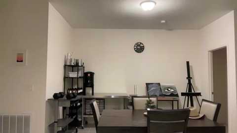 Media: A minimalist, modern studio apartment with white walls, a black wall clock, a black shelf, a desk with a computer, and a tripod.